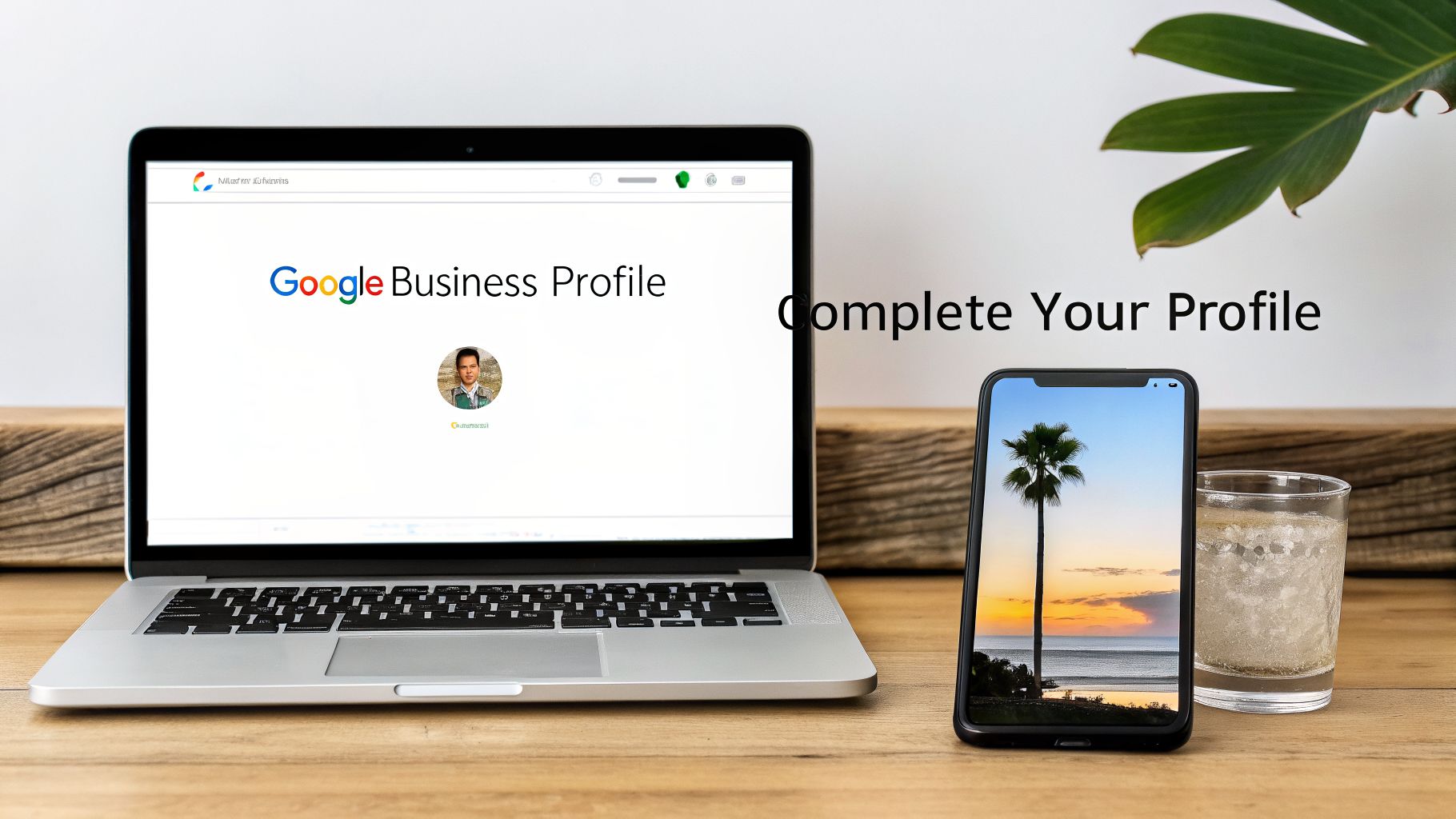 A laptop showing Google Business Profile, a smartphone displaying a beach sunset, and a glass of water on a wooden desk.