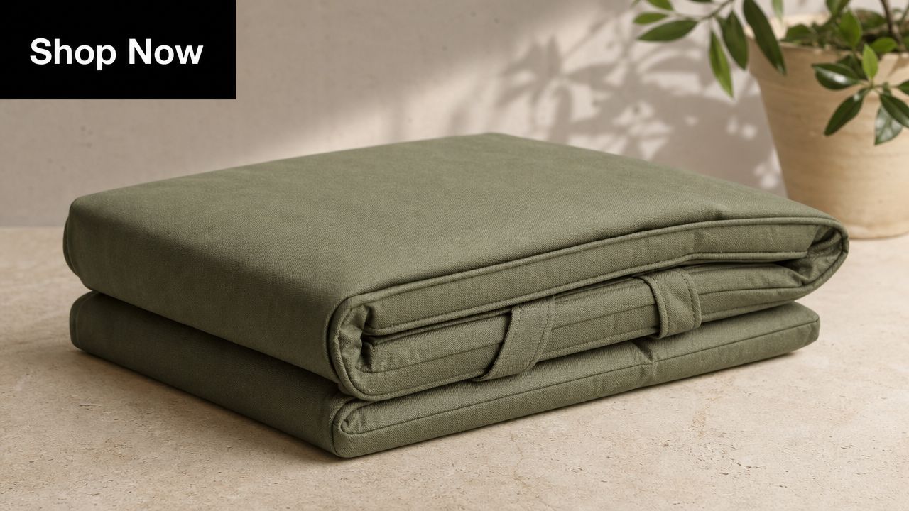 A neatly folded sage green outdoor seat cushion resting on a light beige surface with a plant background.